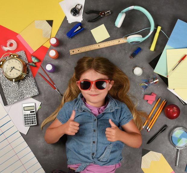 A happy child is holding her thumbs up for a back to school education idea. There are various school supplies around the girl.